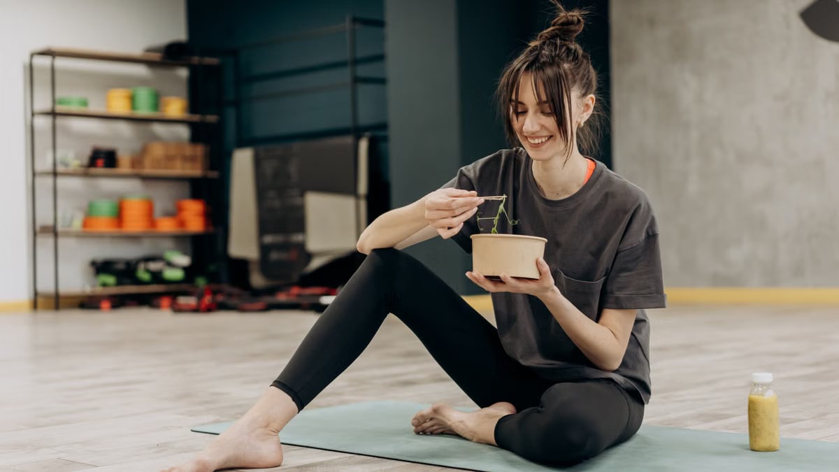 Young woman enjoying a healthy salad after exercising indoors. Promotes a balanced diet and fitness lifestyle.