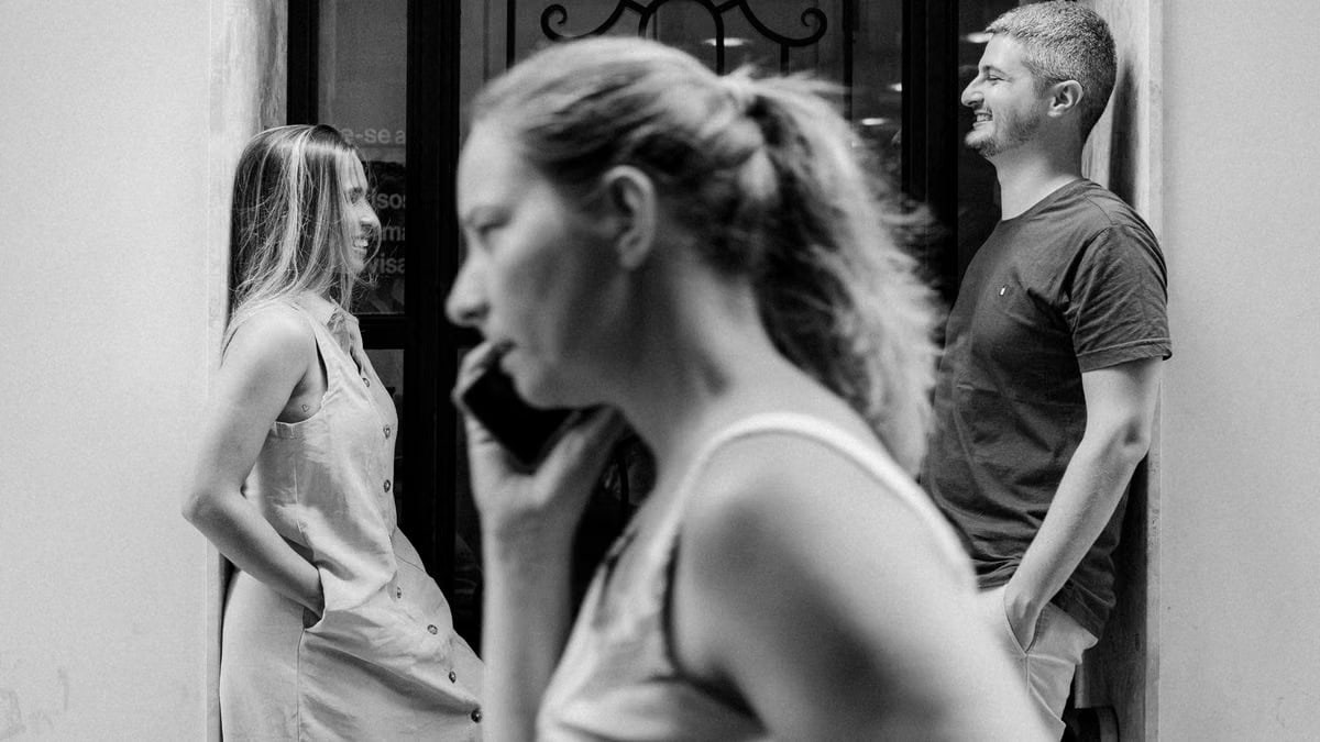 Candid black and white street photo of people interacting and using a phone.